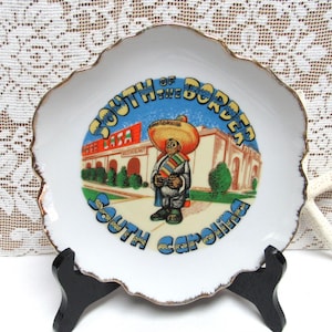 May include: A white ceramic plate with a gold rim and a colorful image of a man in a sombrero standing in front of a building with the text "South of the Border South Carolina".