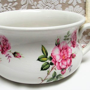 May include: White ceramic chamber pot with a floral design featuring pink roses and green leaves. The pot has a single handle.