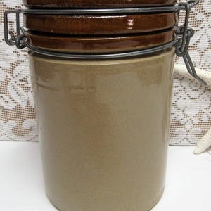 May include: A cylindrical, tan-colored ceramic jar with a dark brown lid and a metal clasp closure. The jar has a smooth, glossy finish and a vintage aesthetic. The lid is composed of three stacked, circular pieces.