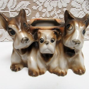 May include: A ceramic planter featuring three dog figurines. The dogs are in shades of brown and white, with detailed facial features. The planter has a rectangular opening for plants.