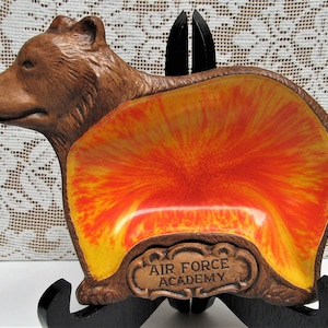 May include: A brown bear-shaped dish with a vibrant orange and yellow interior. The dish has the words "AIR FORCE ACADEMY" in a decorative plaque. The dish is displayed on a black stand, showcasing its unique design.