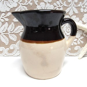 May include: A small, cream-colored ceramic pitcher with a brown and black glaze. The pitcher has a rounded body and a handle.