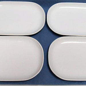 May include: Four white, oval-shaped ceramic plates. The plates have a smooth, glossy finish and are arranged on a blue surface. The plates are a simple, elegant design, suitable for serving food.