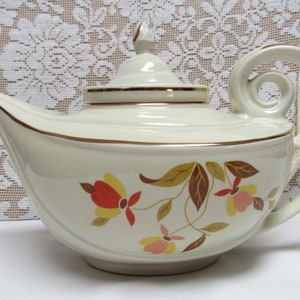 May include: A white teapot with a gold trim and a floral design featuring red, orange, yellow, and pink flowers. The teapot has a curved spout and a handle that curves back towards the body.
