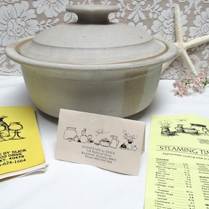 May include: A stoneware casserole dish with a lid, in a light beige color with brown accents. The dish is sitting on a white surface with a yellow card with the text "STONEWARE by SLICK" and a phone number.