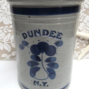 May include: A gray ceramic jar with blue and white painted designs. The jar has the words "Dundee N.Y." on the front.