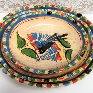 May include: Three nested oval ceramic plates with a hand-painted design of a bird and leaves in shades of blue, green, red, and yellow. The plates have a scalloped edge with alternating green, blue, and red dots.