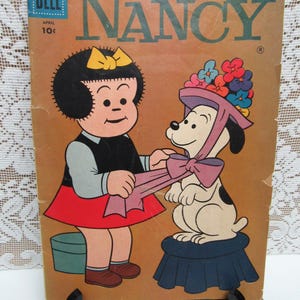 May include: A vintage Dell comic book cover featuring Nancy, a young girl with black hair and a red skirt, tying a pink ribbon on a white dog wearing a flower-adorned hat. The comic book cover is titled "Nancy" and has a price of 10 cents.