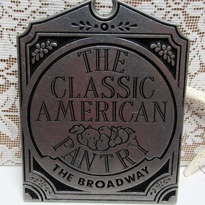 May include: A rectangular, pewter-colored metal sign with the words "THE CLASSIC AMERICAN PANTRY THE BROADWAY" in embossed lettering. The sign has a decorative border with floral accents and a small arched top.