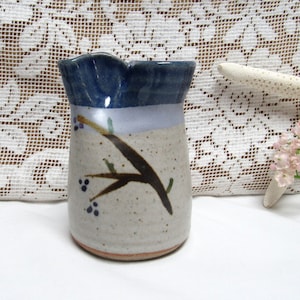 May include: A small, handmade ceramic pitcher with a blue, white, and brown glaze. The pitcher features a painted design of a branch with leaves and berries.
