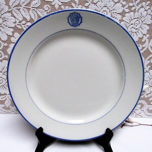 May include: White ceramic plate with a blue rim and a blue decorative emblem. The plate has a thin blue line inside the rim. The plate is displayed on a black stand.