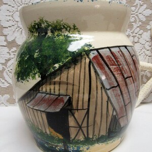 May include: A cream-colored ceramic jar with a hand-painted scene of a rustic wooden barn featuring a rusty metal roof, a large green tree, and blue floral accents at the base. The jar has a wide opening and a matching lid with blue speckles.