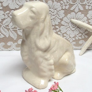 May include: Cream-colored ceramic dog figurine in a seated position. The dog has long, floppy ears and a detailed, textured surface. The figurine is a decorative item, possibly a collectible or home decor piece.