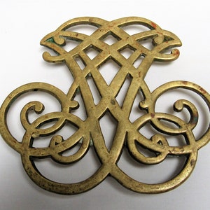 May include: A brass trivet with an intricate design featuring a woven pattern and swirls. The trivet is a light gold color and has a worn, antique look.