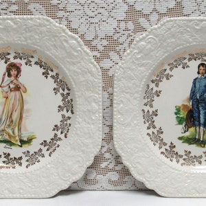 May include: Two white decorative plates with scalloped edges. Each plate features a central image of a person in period clothing, surrounded by a gold floral border. The plates are on a white lace tablecloth.