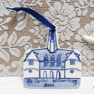 May include: A white ceramic ornament with a blue illustration of a building, featuring a tall steeple and arched windows. The text "The Capitol Williamsburg 2001" is written in blue. A blue ribbon is attached for hanging.