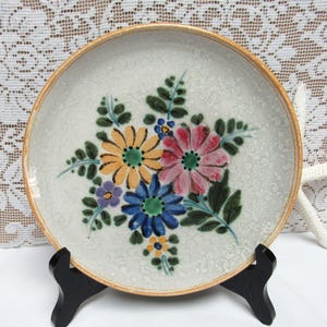 May include: A white ceramic plate with a floral design. The flowers are painted in shades of yellow, pink, blue, and green. The plate has a speckled glaze and a brown rim.