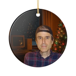 May include: Round Christmas ornament featuring a man wearing a baseball cap and plaid shirt. The background includes a Christmas tree, a computer, and a starry night scene. The ornament is suspended by a gold string.