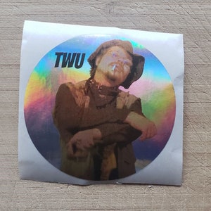 May include: A round sticker with a holographic finish featuring a man wearing a hat and a brown jacket. The sticker has the text "TWU" printed in black.