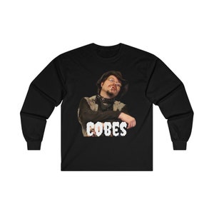 May include: Black long-sleeve t-shirt with a white graphic of a man wearing a cowboy hat and a bandana. The text "COBES" is printed in white below the image.
