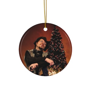 May include: Round Christmas ornament featuring a person in a hat and glasses, with a lit cigarette, posing in front of a Christmas tree. The ornament has a gold string for hanging.