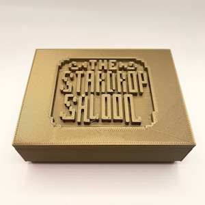 May include: A gold-colored rectangular box with the words "THE STARDROP SALOON" embossed on the lid. The text is in a stylized, retro font, and the box has a textured surface.