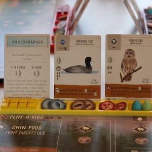 May include: A board game setup featuring cards with bird illustrations and text. The cards include "Photographer," "American Coot," and "Barred Owl." Tokens in various colors are in a yellow tray. The game board is visible in the background.