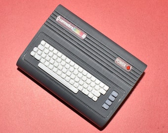 Peut inclure: Un ordinateur personnel Commodore 16 noir avec un clavier blanc et un bouton d'alimentation rouge. L'ordinateur a un logo noir et arc-en-ciel qui dit "Commodore 16".