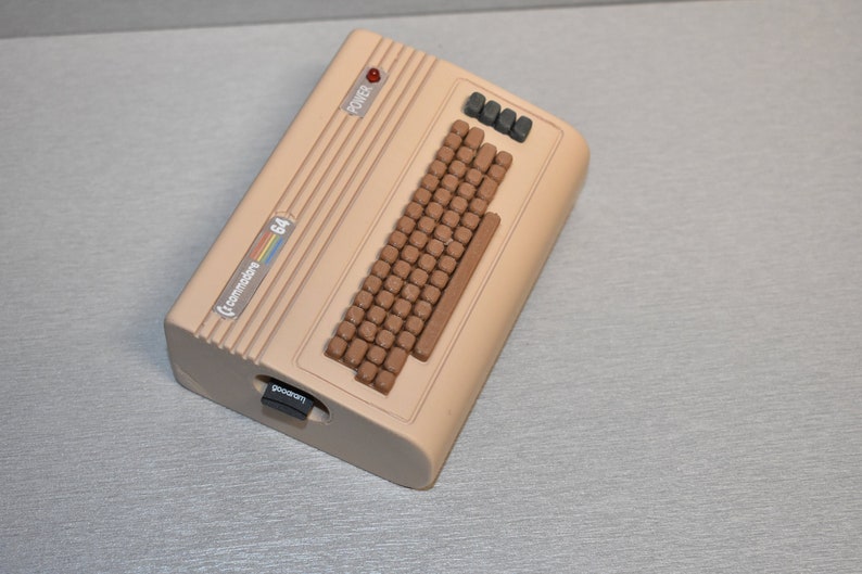Peut inclure: Un ordinateur Commodore 64 beige avec un clavier brun et un voyant d'alimentation rouge. L'ordinateur poss&egrave;de un logement de cartouche &eacute;tiquet&eacute; "gameport" et un autocollant indiquant "Commodore 64".
