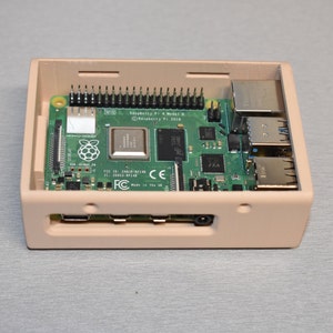 Peut inclure: Un bo&icirc;tier en plastique beige avec une carte de circuit imprim&eacute; verte &agrave; l'int&eacute;rieur. La carte de circuit imprim&eacute; porte l'inscription "Raspberry Pi 4 Model B" et "Raspberry Pi 2018". Le bo&icirc;tier poss&egrave;de plusieurs ports &agrave; l'avant, notamment des ports USB et HDMI.