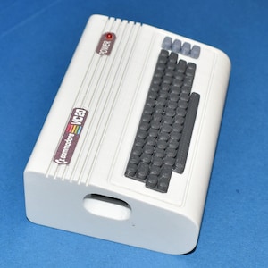 Può includere: Un computer Commodore VIC-20 bianco con una tastiera grigia e un pulsante di accensione rosso. Il computer ha un logo arcobaleno sul lato.