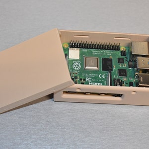 Peut inclure: Un bo&icirc;tier en plastique beige avec un circuit imprim&eacute; vert &agrave; l'int&eacute;rieur. Le circuit imprim&eacute; porte l'inscription "Raspberry Pi 4 Model B" et "Raspberry Pi 3B+". Le bo&icirc;tier est ouvert et le circuit imprim&eacute; est visible.