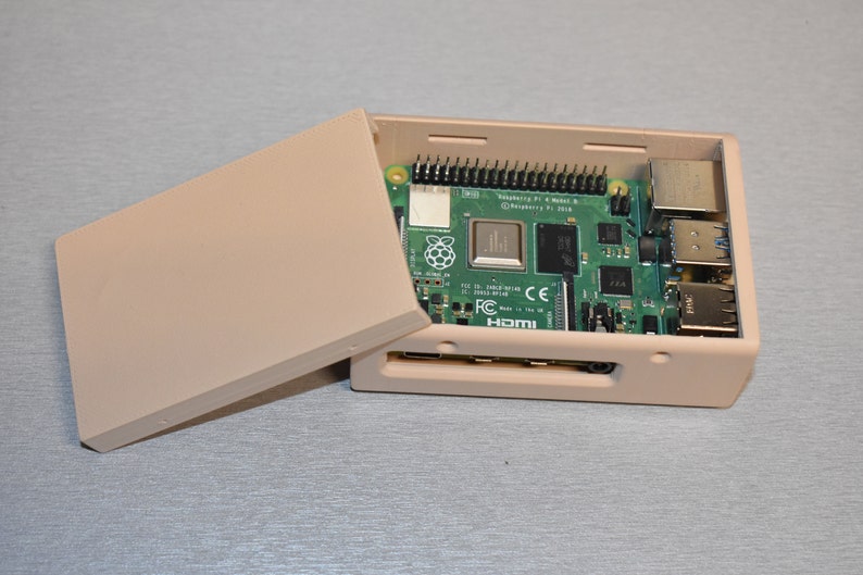 Peut inclure: Un bo&icirc;tier en plastique beige avec un circuit imprim&eacute; vert &agrave; l'int&eacute;rieur. Le circuit imprim&eacute; porte l'inscription "Raspberry Pi 4 Model B" et "Raspberry Pi 3B+". Le bo&icirc;tier est ouvert et le circuit imprim&eacute; est visible.