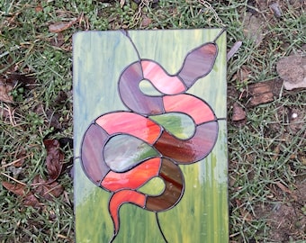 May include: A stained glass window panel featuring a red, brown and pink snake on a green background.