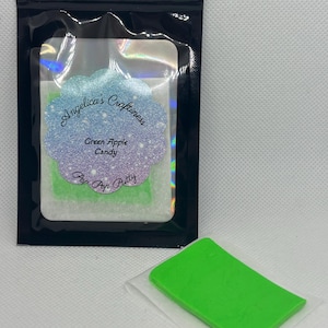 May include: A black, shiny pouch containing a clear package with green apple candy. The clear package has a label with the text "Angelica's Craftiness" and "Green Apple Candy". A separate, bright green candy is on a white paper.