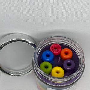 May include: A glass jar candle with a purple wax base and a rainbow label. The label reads "Aria's Candles Fruit Loops". The candle is topped with six colorful donut-shaped wax pieces.