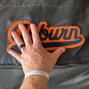 Puede incluir: Parche de fieltro naranja y azul marino con la palabra "Auburn" en fuente cursiva. El parche tiene la forma de la palabra y tiene un contorno azul marino. El fondo es gris oscuro.