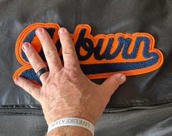 May include: An orange and navy blue felt patch with the word "Auburn" in a script font. The patch is in the shape of the word and has a navy blue outline. The background is a dark gray.