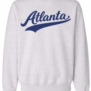 以下が含まれることがあります： 「Atlanta」の文字が紺色の筆記体でプリントされたライトグレーのスウェットシャツ。文字にはスタイリッシュな下線が入っています。スウェットシャツはクルーネックで長袖です。
