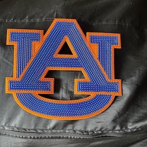 May include: A blue and orange embroidered patch featuring the letter "A" and the word "U" in a stylized design. The patch has a textured appearance and is set against a black background.