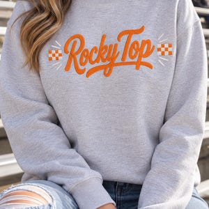 Pode incluir: Uma camisola de gola redonda cinzenta clara com o texto Rocky Top bordado a laranja em letra cursiva. O design inclui pequenos quadrados axadrezados a laranja e branco em ambos os lados do texto, acentuados por linhas bordadas finas.