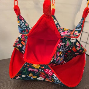 Puede incluir: Una peque&ntilde;a hamaca para mascotas con interior rojo y exterior verde oscuro estampado con flores, setas y mariposas de colores. La hamaca est&aacute; suspendida por clips de pl&aacute;stico rojos y amarillos.