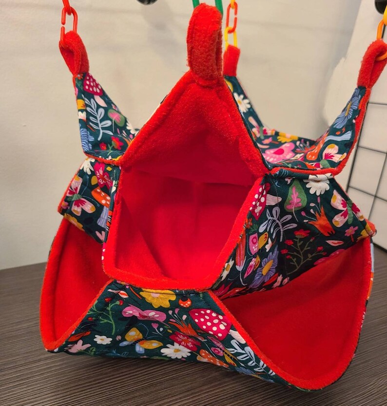 Puede incluir: Una peque&ntilde;a hamaca para mascotas con interior rojo y exterior verde oscuro estampado con flores, setas y mariposas de colores. La hamaca est&aacute; suspendida por clips de pl&aacute;stico rojos y amarillos.