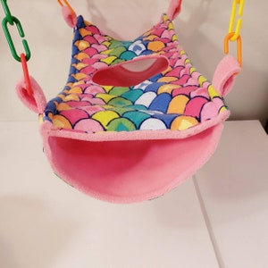 May include: A colorful pet hammock with a mermaid scale pattern in shades of pink, blue, yellow, and green. The hammock has a pink interior and is suspended by plastic chains.