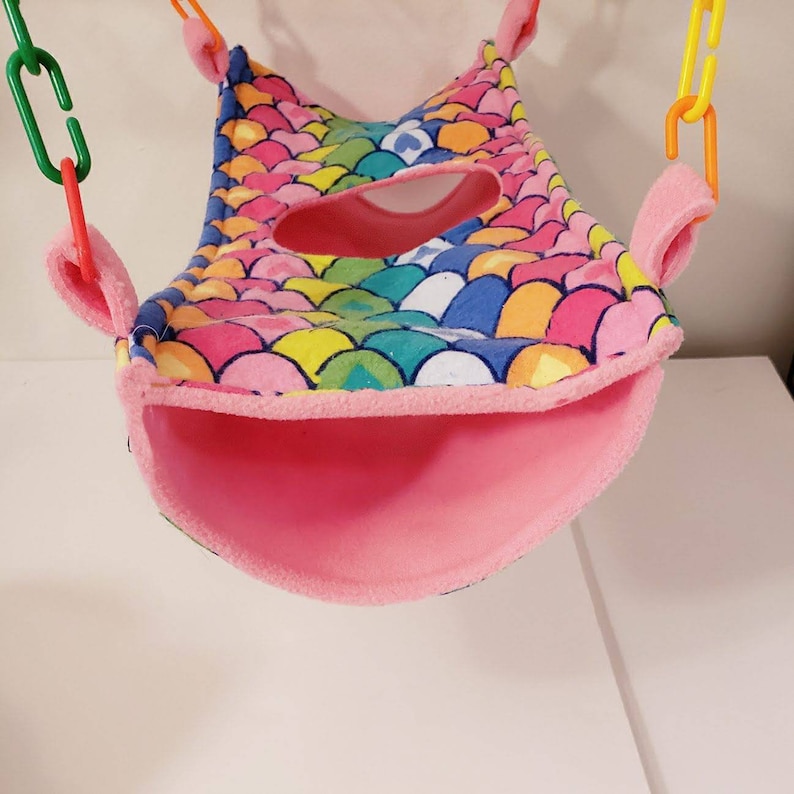 May include: A colorful pet hammock with a mermaid scale pattern in shades of pink, blue, yellow, and green. The hammock has a pink interior and is suspended by plastic chains.