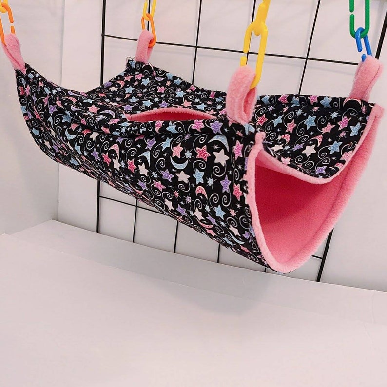 May include: A small pet hammock with a black fabric exterior patterned with colorful stars and moons. The interior is soft pink fleece. The hammock is suspended by yellow and orange plastic chain links.