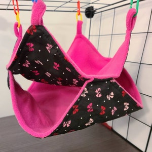 May include: A pink and black pet hammock. The hammock is made of soft fleece material. The black fabric is patterned with pink and red bows and white polka dots. The hammock is suspended by colorful plastic clips.
