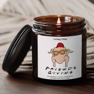 May include: A lit soy wax candle in a brown glass jar with a black lid. The label features a cartoon turkey wearing sunglasses and a chef's hat, with the text "FRIENDS GIVING". The candle is eco-friendly and made in the USA.