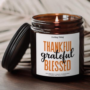 May include: A lit brown glass jar candle with a black lid. The label reads "THANKFUL grateful BLESSED" in orange and black. The candle is made with natural soy wax.