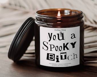 May include: A lit candle in a brown glass jar with a black lid. The label reads "You a Spooky Bitch" in a bold, cut-out style font. The candle is labeled "Feeling Thing" and is made with natural soy wax.
